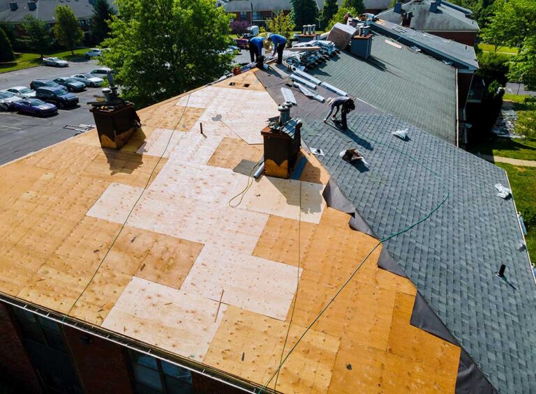 Roofing worker rolls synthetic underlayment across a plywood roof deck beneath shingles.